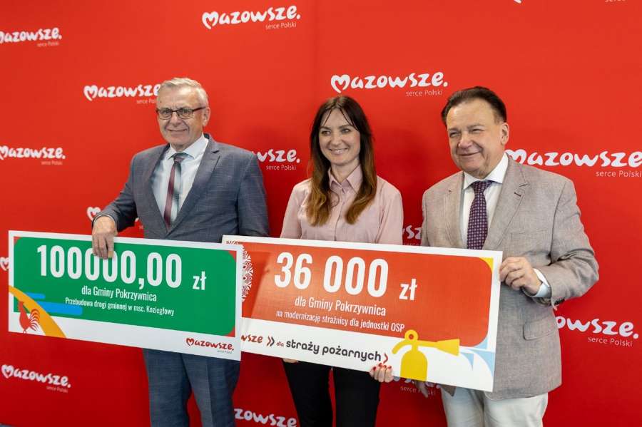 Adam Strucik i dwie osoby trzymają dwa pamiątkowe czeki dotacji dla Gminy Pokrzywnica na tle czerwonej ścianki z logo Mazowsze.